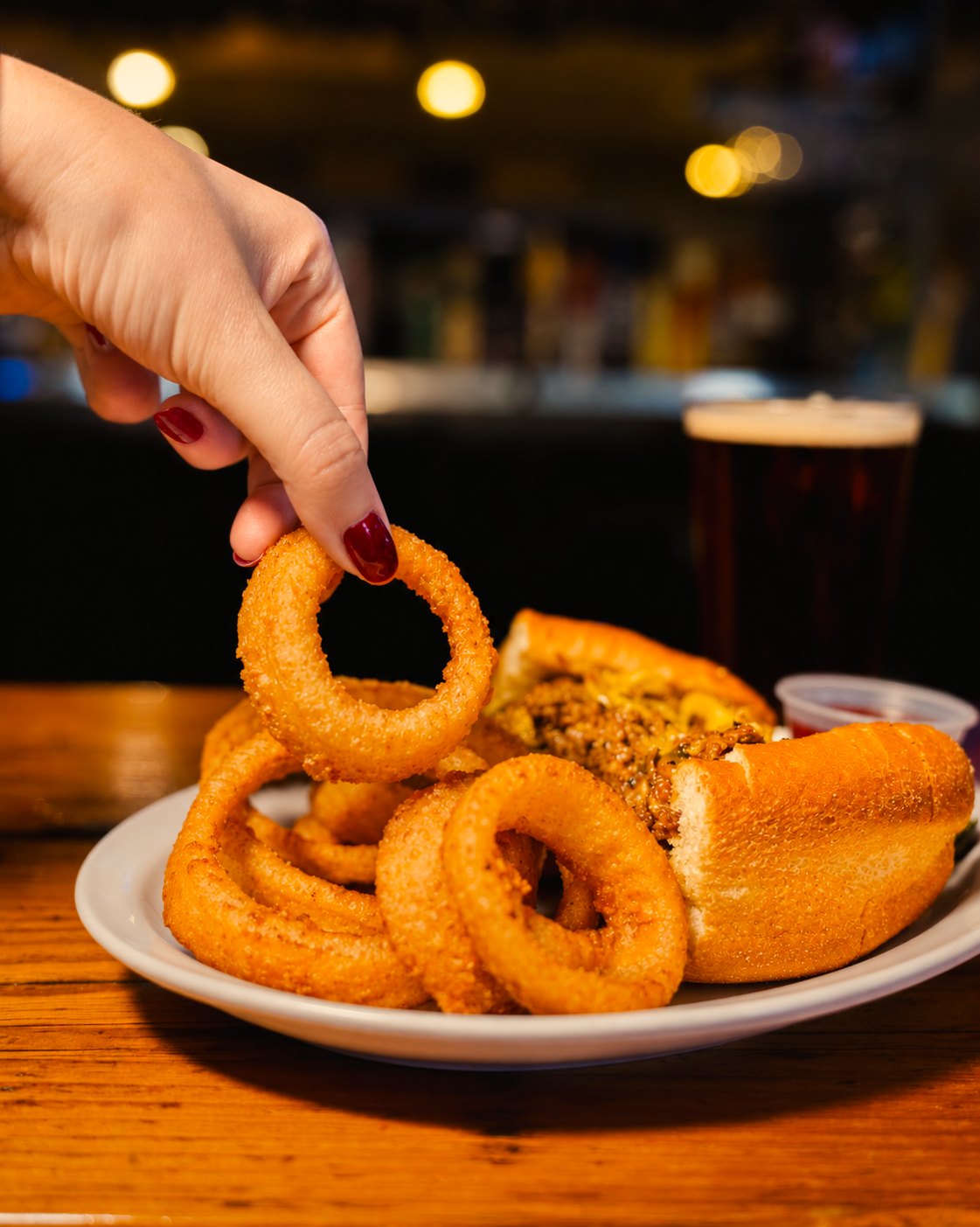 Hand picking up a crispy onion ring from a plate with a sandwich and a pint